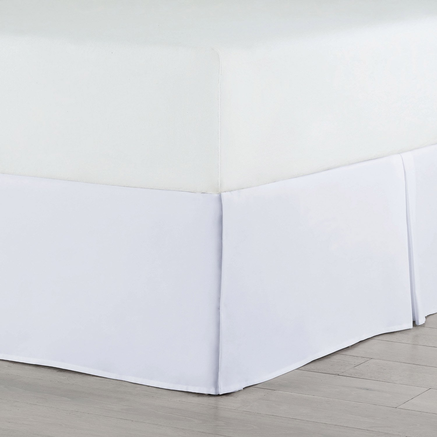 A close-up of a bed corner shows the Martex Basics Bedskirt by WestPoint Home in crisp white, neatly draping to the floor over a plain mattress, resembling hotel bedding and placed on a light wood floor.