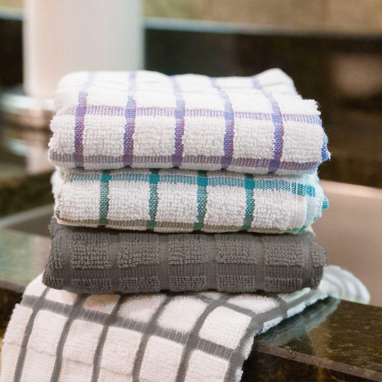 Four Martex Kitchen Collection Windowpane kitchen towels by WestPoint Home, in purple, teal, and gray checkered patterns, are stacked and draped on a countertop near the kitchen sink, adding style to the space.
