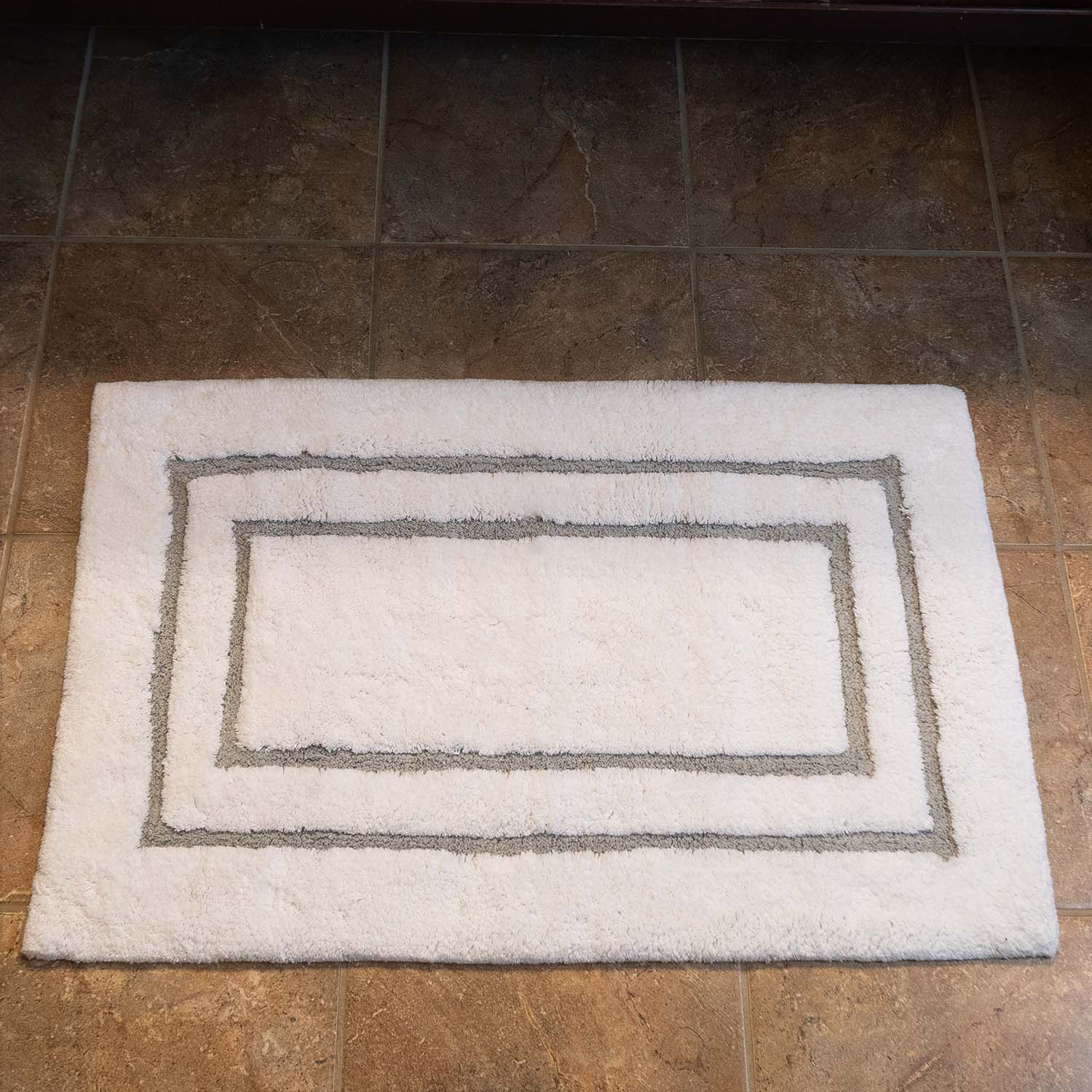 A rectangular white Gryphon Collection - Tufted Bath Rug by WestPoint Home with two gray borders rests on a brown tiled floor.
