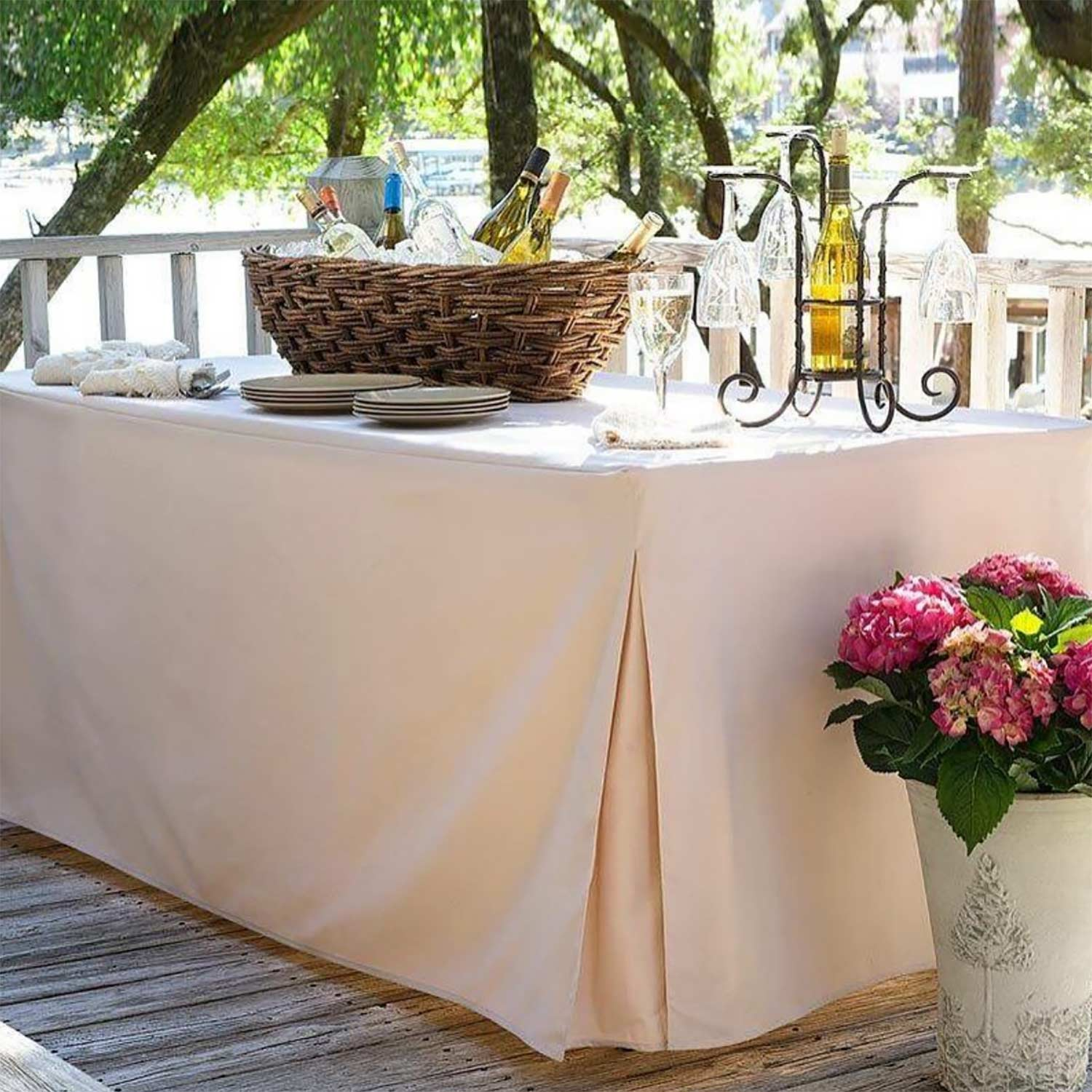 A wooden deck among trees features an outdoor table with the WestPoint Home Solids - Platinum - Table Cover in White, holding wine bottles in a wicker basket, wine glasses on a metal rack, plates, napkins, and pink flowers in a white pot nearby.