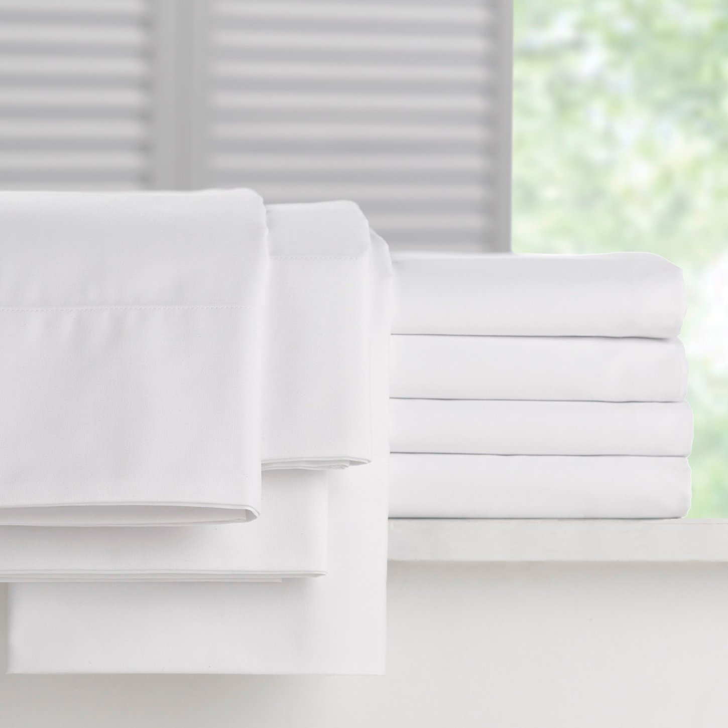 A neatly folded stack of WestPoint Home Martex Millennium 250 Bone sheets and pillow cases rests on a white surface, with a softly blurred background of a window and greenery outside.