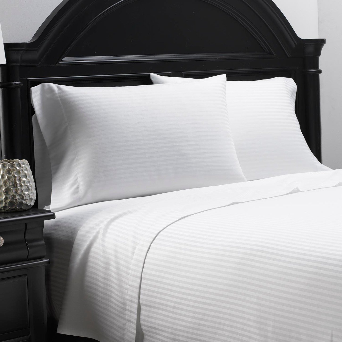 A neatly made bed features WestPoint Home’s Martex DryFast 200 Fitted Sheet with a black arched headboard and eco-friendly white bedding, complemented by two pillows and a black nightstand with a decorative lamp.