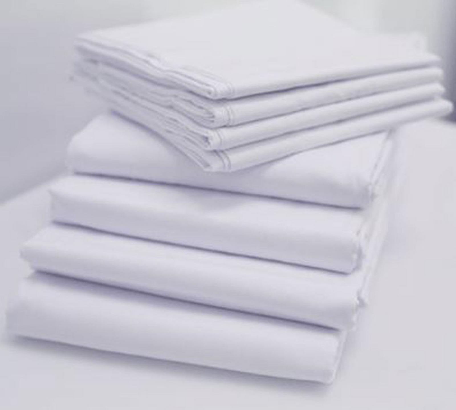 A neatly stacked pile of 1Concier Flat Sheet Solid Percale Blend bed sheets rests on a white surface, four on the bottom and three on top. A softly blurred background highlights their clean, orderly arrangement.