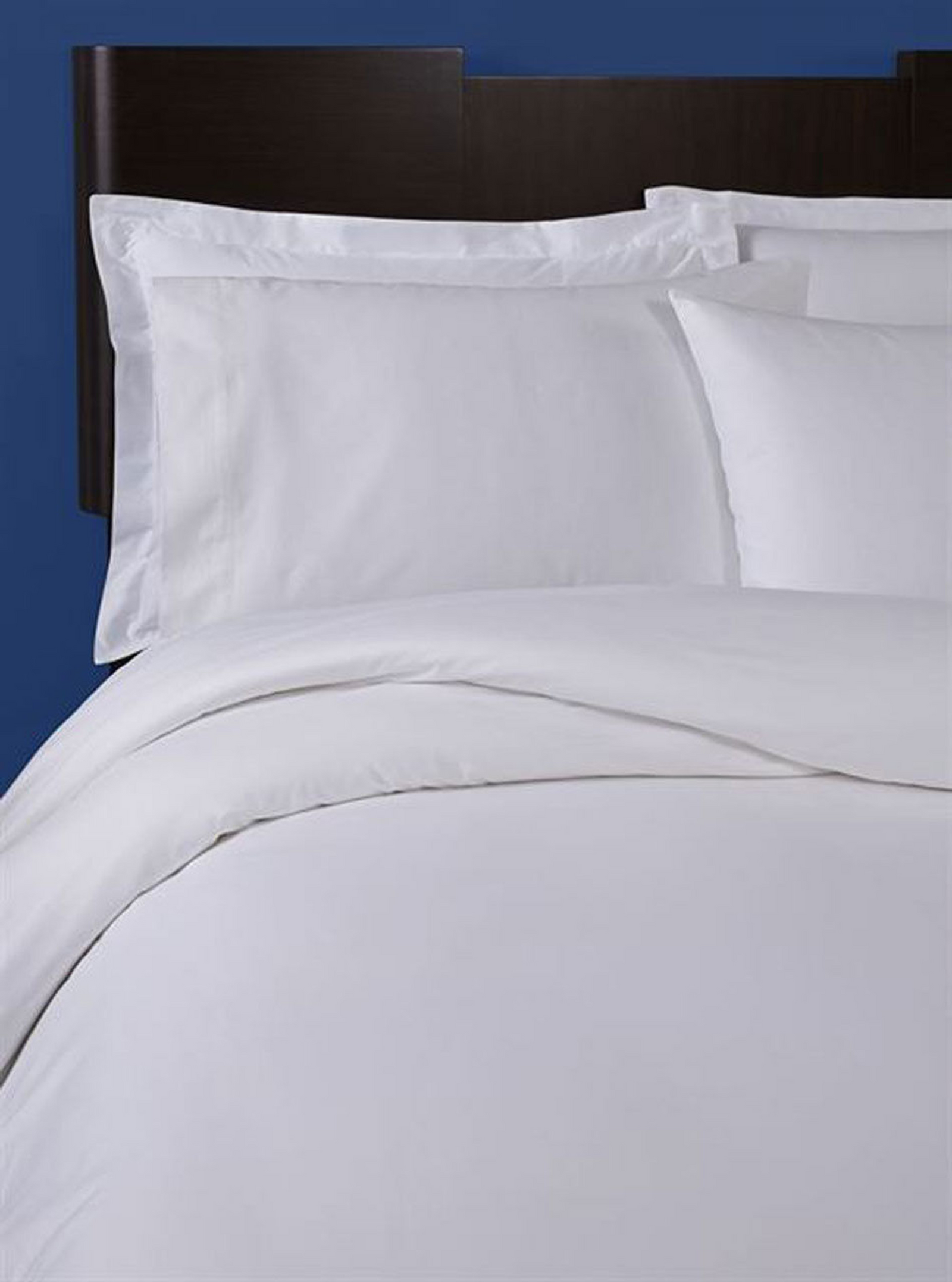 A neatly made bed with 1Concier's Duvet Cover Percale T300 and pillows, set against a dark wooden headboard and blue wall. The percale bedding from 1Concier looks soft and inviting.