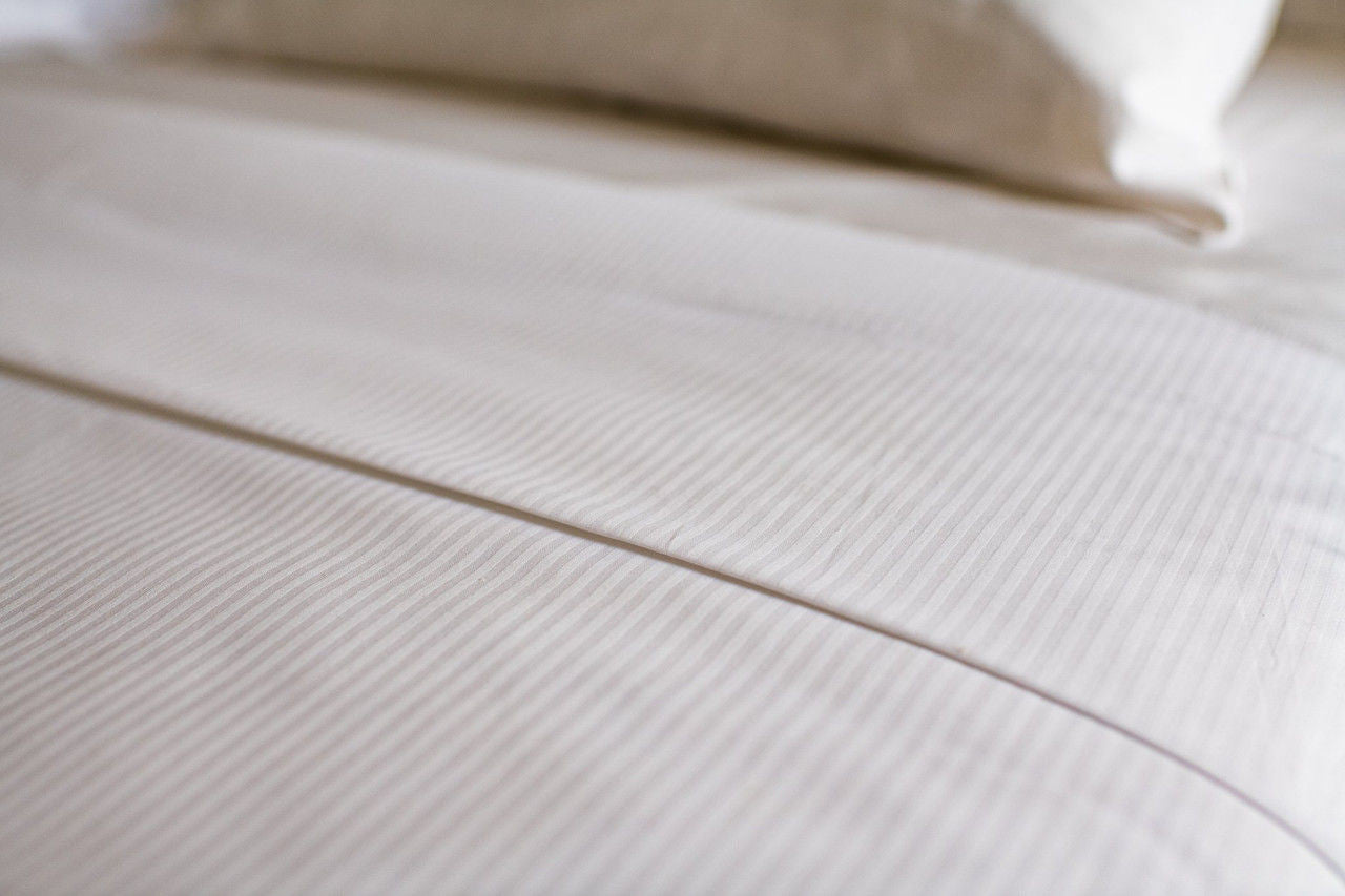 A close-up of a neatly made bed with Thomaston Mills Narrow Stripe 250 Thread Count Satin Stripe sheets, crafted from a soft combed cotton blend and featuring luxurious triple sheeting for added comfort.