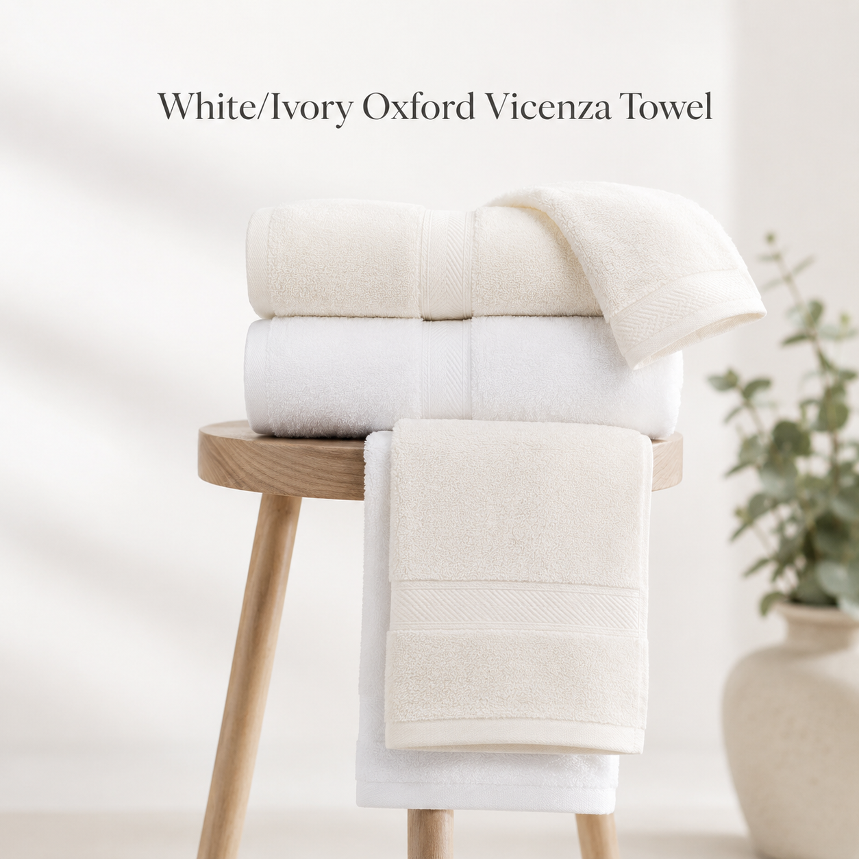 A White/Ivory Oxford Vicenza Bath Sheet / Pool Towel by Ganesh Mills rests folded on a wooden stool, with a blurred green plant in the background. Text reads: White/Ivory Oxford Vicenza ringspun cotton towel.
