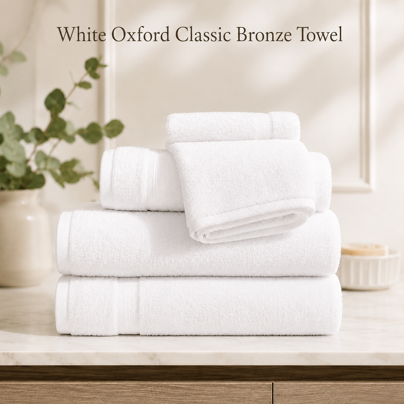 A stack of white Ganesh Mills Bath Towel - White Oxford Classic Bronze Towels is neatly arranged on a marble countertop, with a plant and beige vase in the background. Text above reads: Perfect for quality wholesale towels seekers.