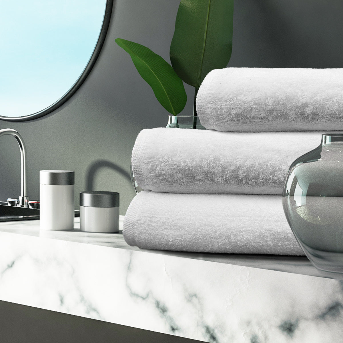 Three neatly folded Ganesh Mills Hand Towel - White Oxford Platinum Towels rest on a marble countertop beside a vase, containers, and mirror—ideal for those seeking luxury or bulk hotel towels in a modern bathroom setting.