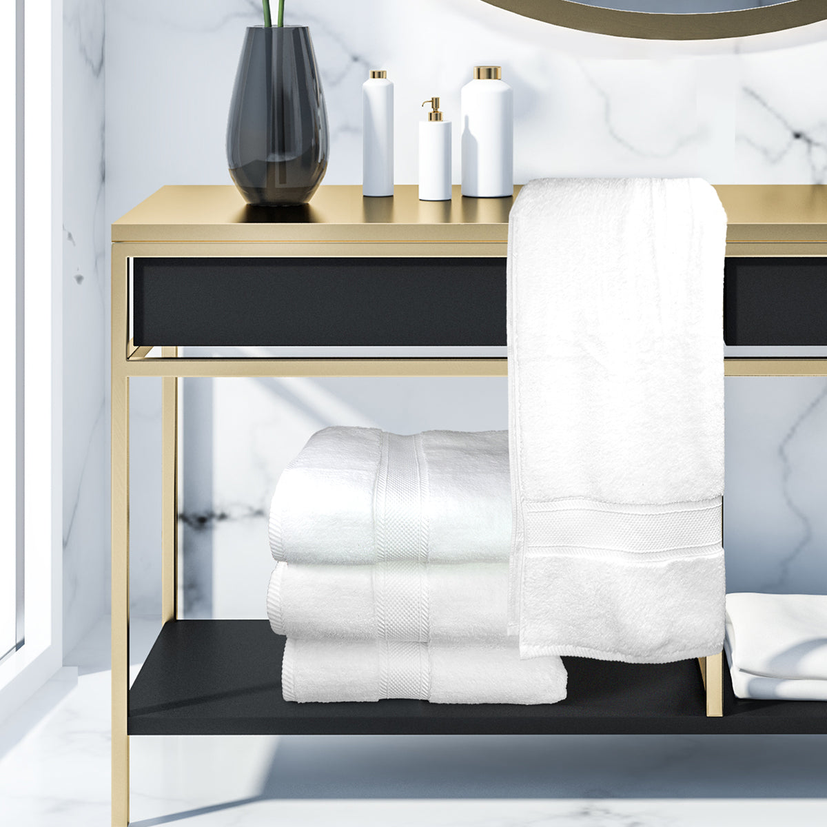 A modern bathroom vanity holds a black vase, white bottles, and luxury hotel towels—including the Ganesh Mills Washcloth - White Oxford Miasma Towel—folded and draped over the edge, all set against a marble wall background.