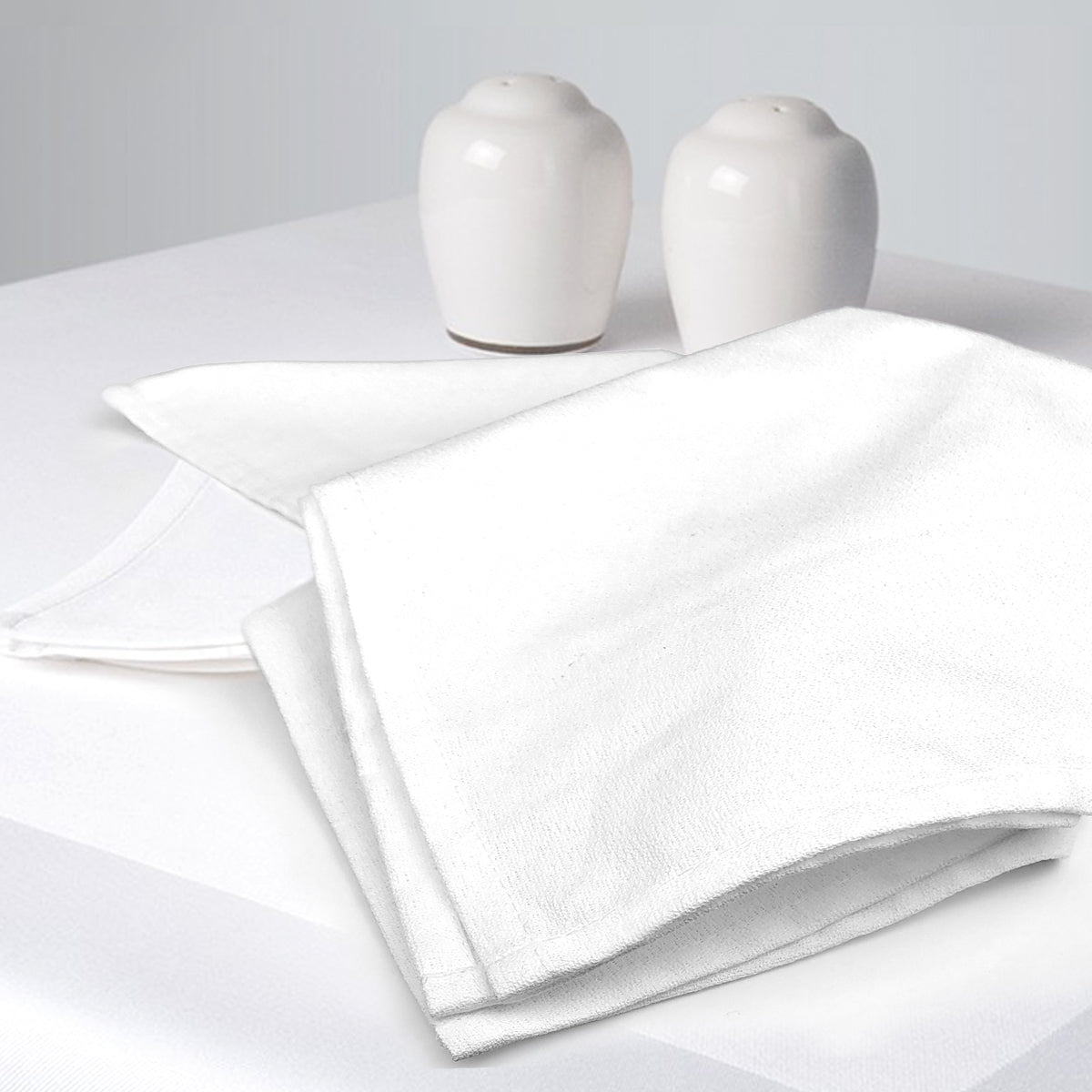 Two white cloth napkins are neatly folded on a Ganesh Mills Tablecloth - White Momie Banquet Table Linen, with white ceramic salt and pepper shakers placed in the background.