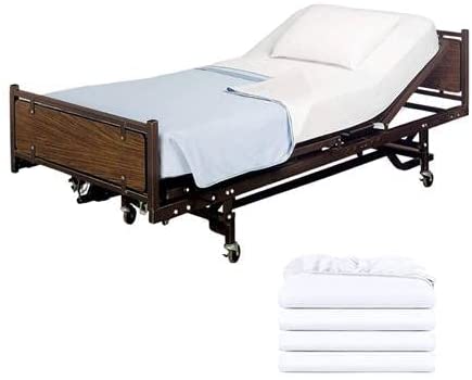 A hospital bed with an adjustable head section, white mattress, pillow, and light blue blanket stands beside a neatly folded stack of KSE Knitted Fitted Sheets—perfect for keeping hospital beds fresh and clean.