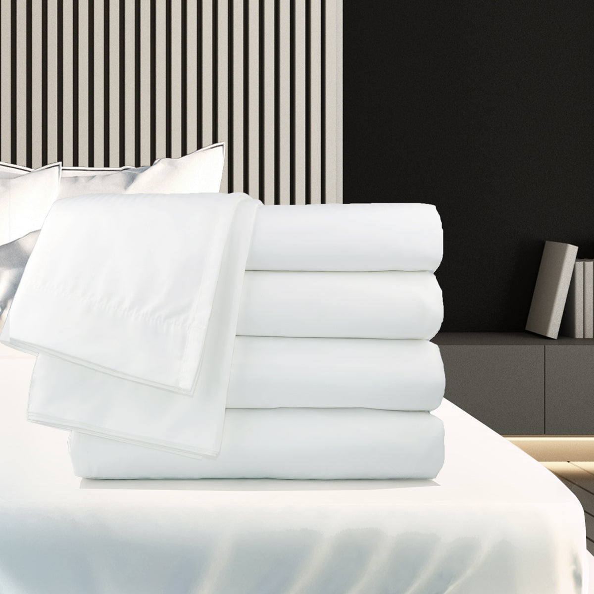 A neatly folded stack of Ganesh Mills Oxford T200 Superblend Flat Sheets with 2" top hem and 1" bottom hem is displayed on a bed in a modern bedroom featuring striped wall panels and decorative pillows.