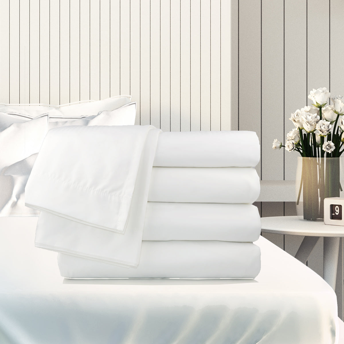 A stack of Ganesh Mills Oxford Super T300 Bed Linen Fitted Sheets sits on a white bed in a bright room, with a nightstand holding white flowers and a digital clock against a paneled wall.