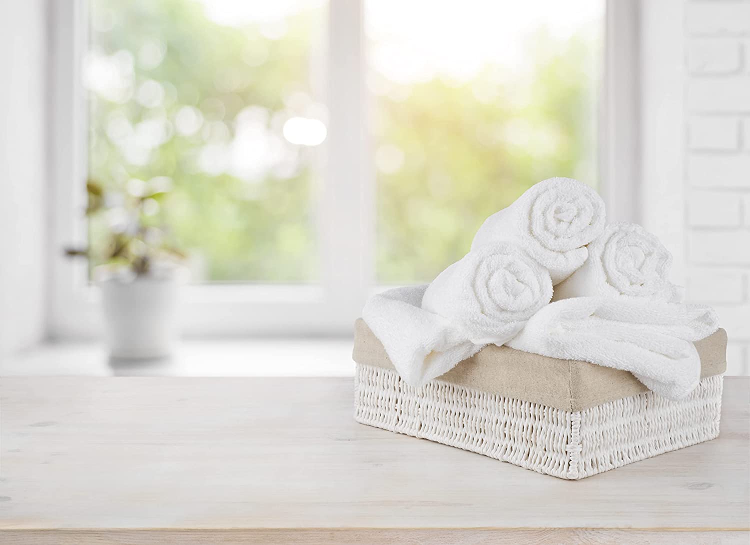 A wicker basket filled with KSE Bath Towel - Standard 16 Single Towels sits on a light wooden surface, with a bright window and a potted plant blurred in the background.