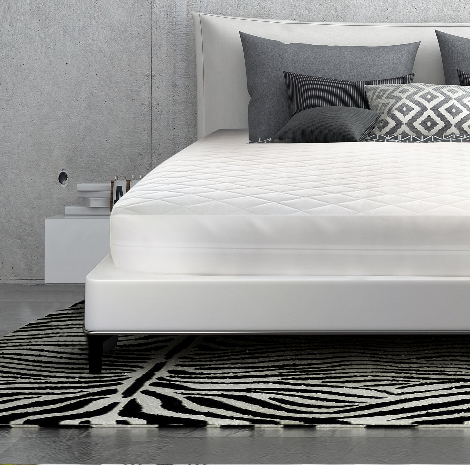 A modern bed with a white mattress topped by the Oxford Three Layer Quilted Bed Pad Waterproof Fitted from Ganesh Mills, gray and patterned pillows, and a geometric black-and-white rug, set against a concrete wall with a white nightstand on the left.