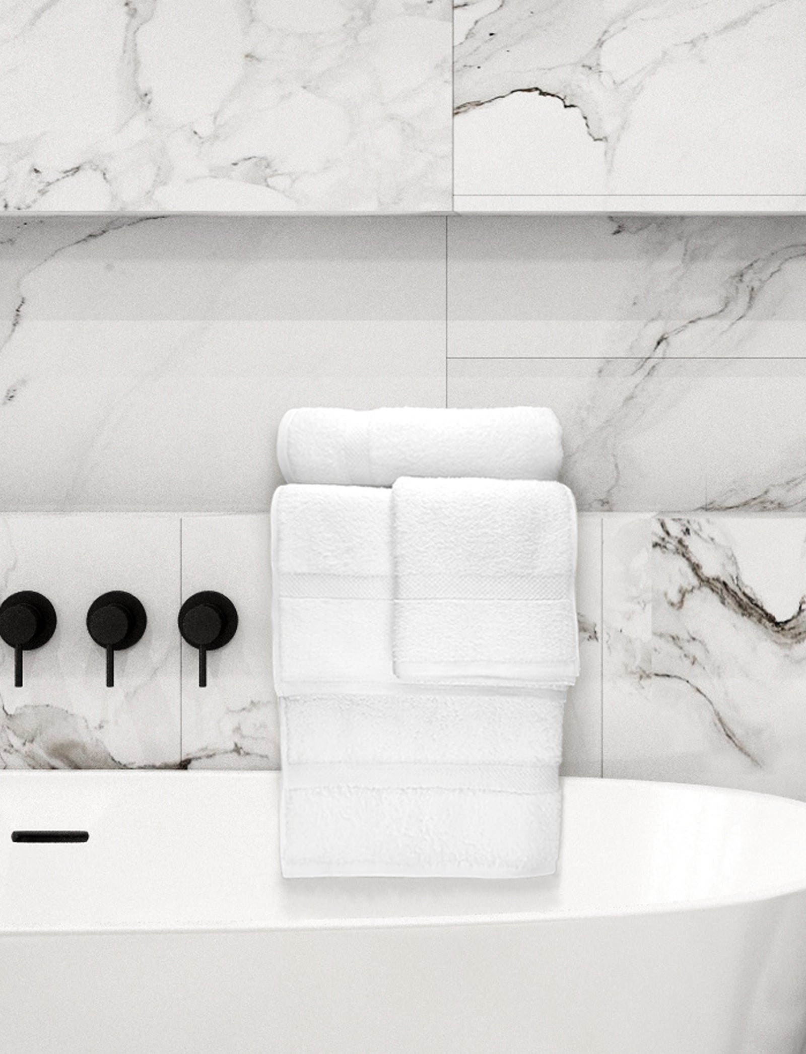 A stack of Ganesh Mills Washcloth - White Oxford Nuvola Towels is neatly arranged on the edge of a white bathtub, set against white marble tiles with gray veining and matte black faucet handles.